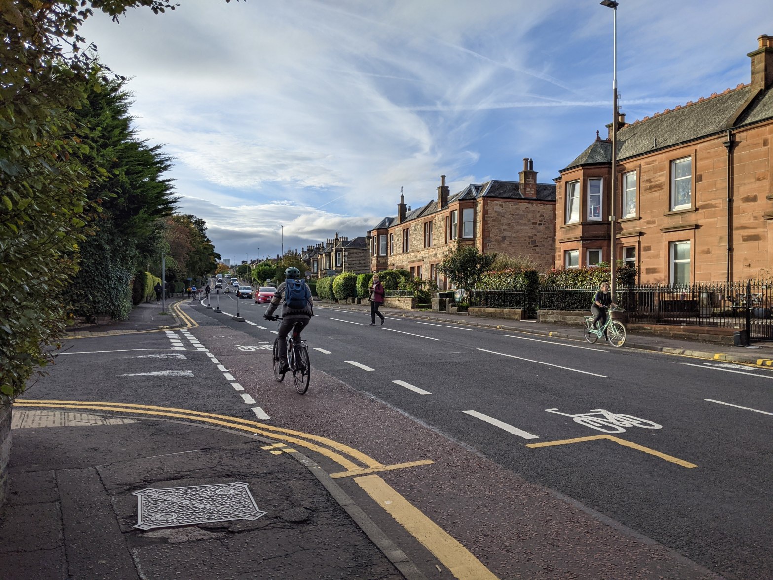 Photograph of a street with cycle lanes painted on either side. Cyclists can be seen on either side of the street. A pedestrian is crossing the road, looking unhurried. Trees are on the left hand side of the street; houses in honey coloured stone on the right.